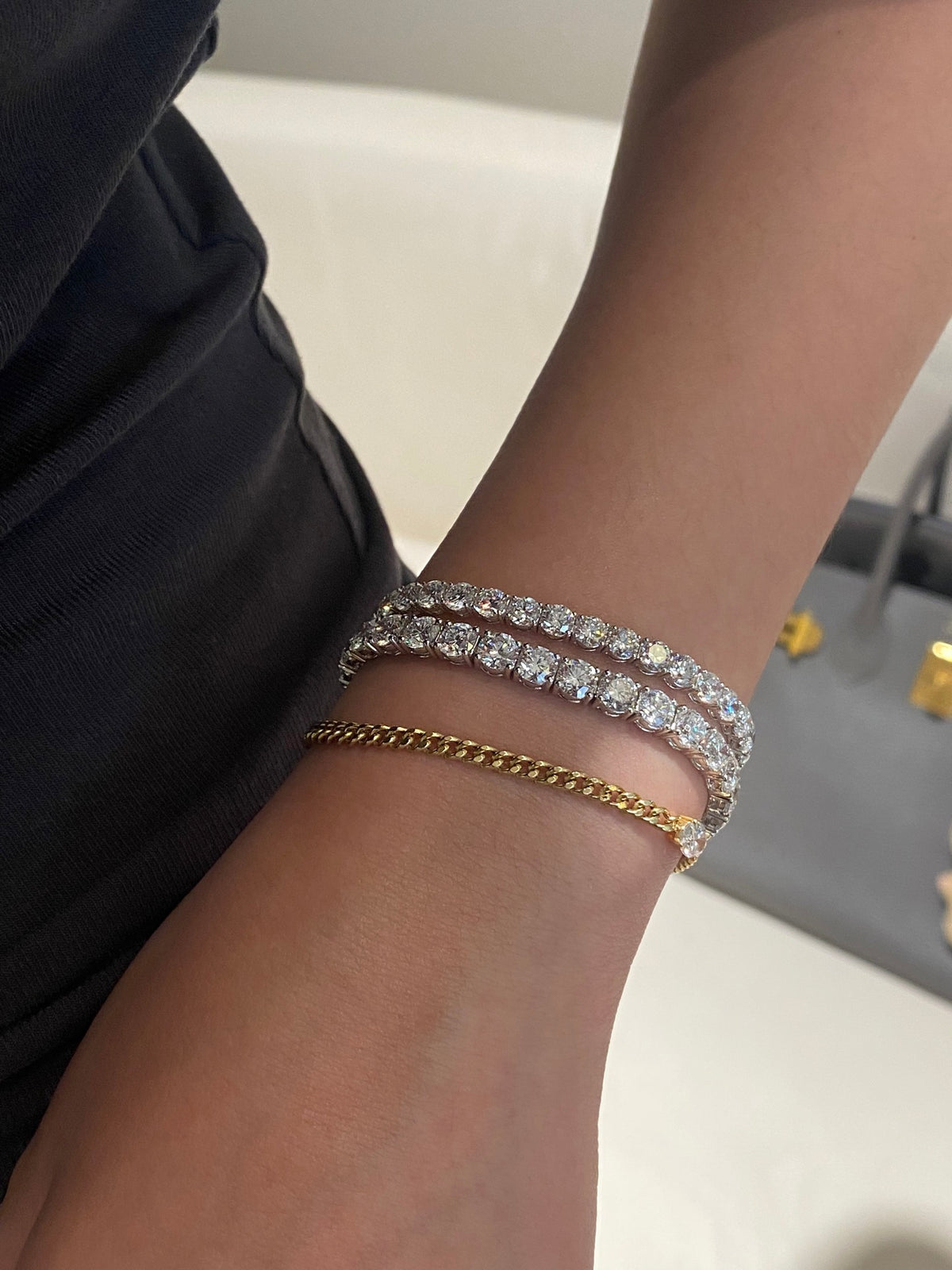 Larger white-metal round diamond tennis bracelet nearest the hand, shown with a smaller tennis bracelet and separate gold chain bracelet